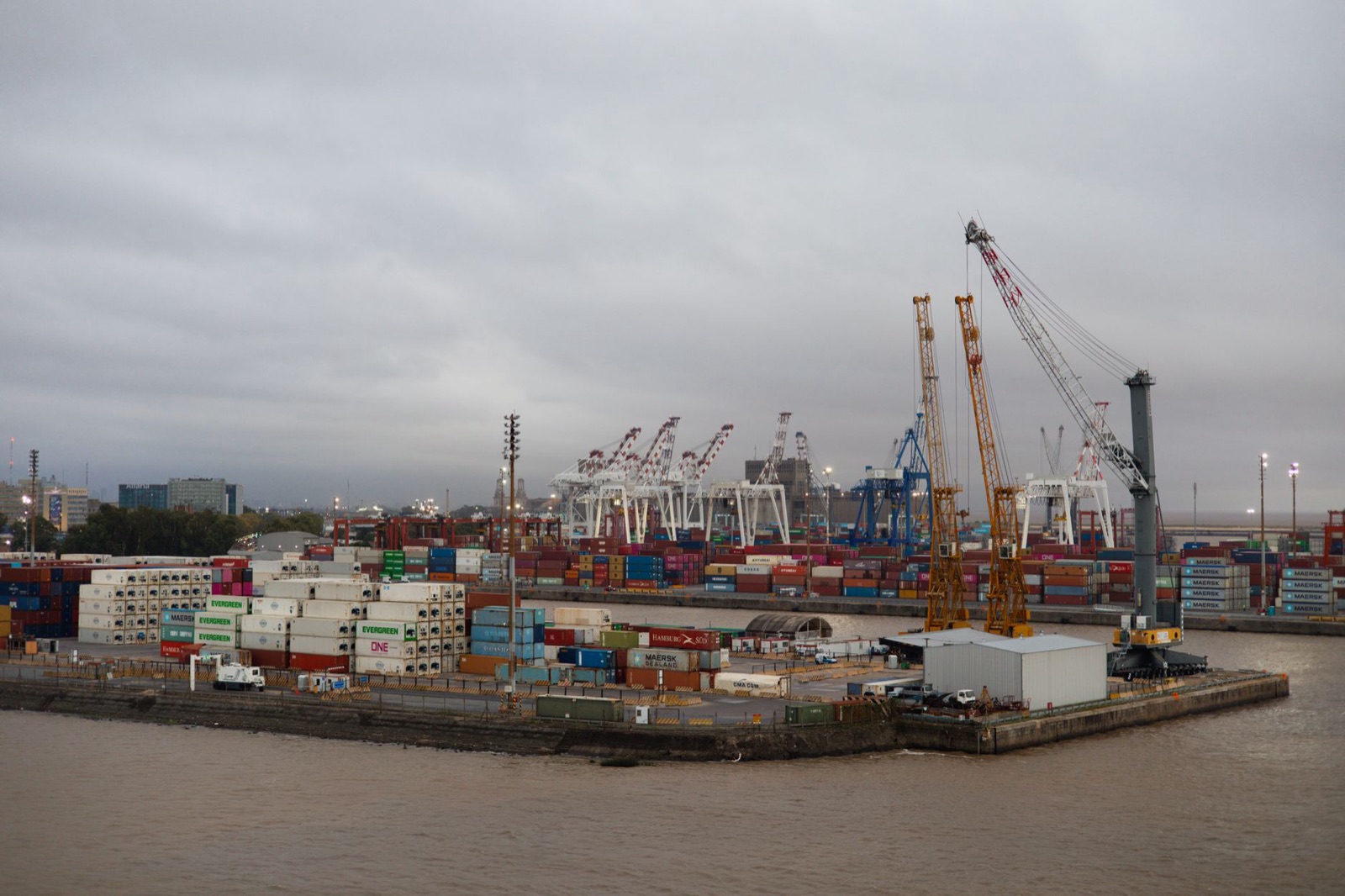 Expansive view of a busy industrial port with shipping containers and cranes under cloudy skies.